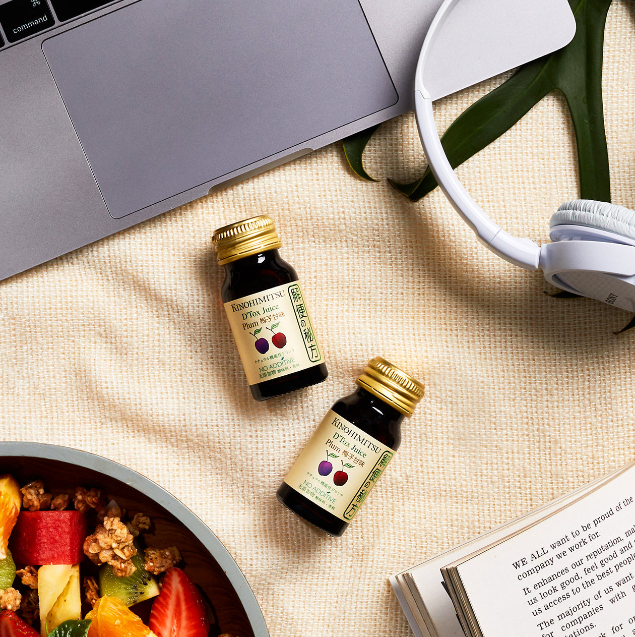 Two bottles of Kinohimitsu D'tox Plum Juice placed on a beige fabric beside a laptop, headphones, and a bowl of fresh fruit and granola, highlighting a healthy lifestyle setting.